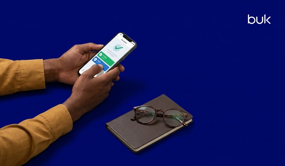 Manos usando un celular para el control de asistencia de personal, junto a una libreta y lentes sobre fondo azul.