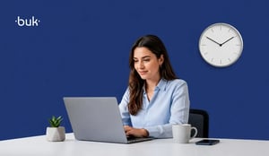 Mujer profesionista en escritorio con laptop con reloj de pared al fondo.